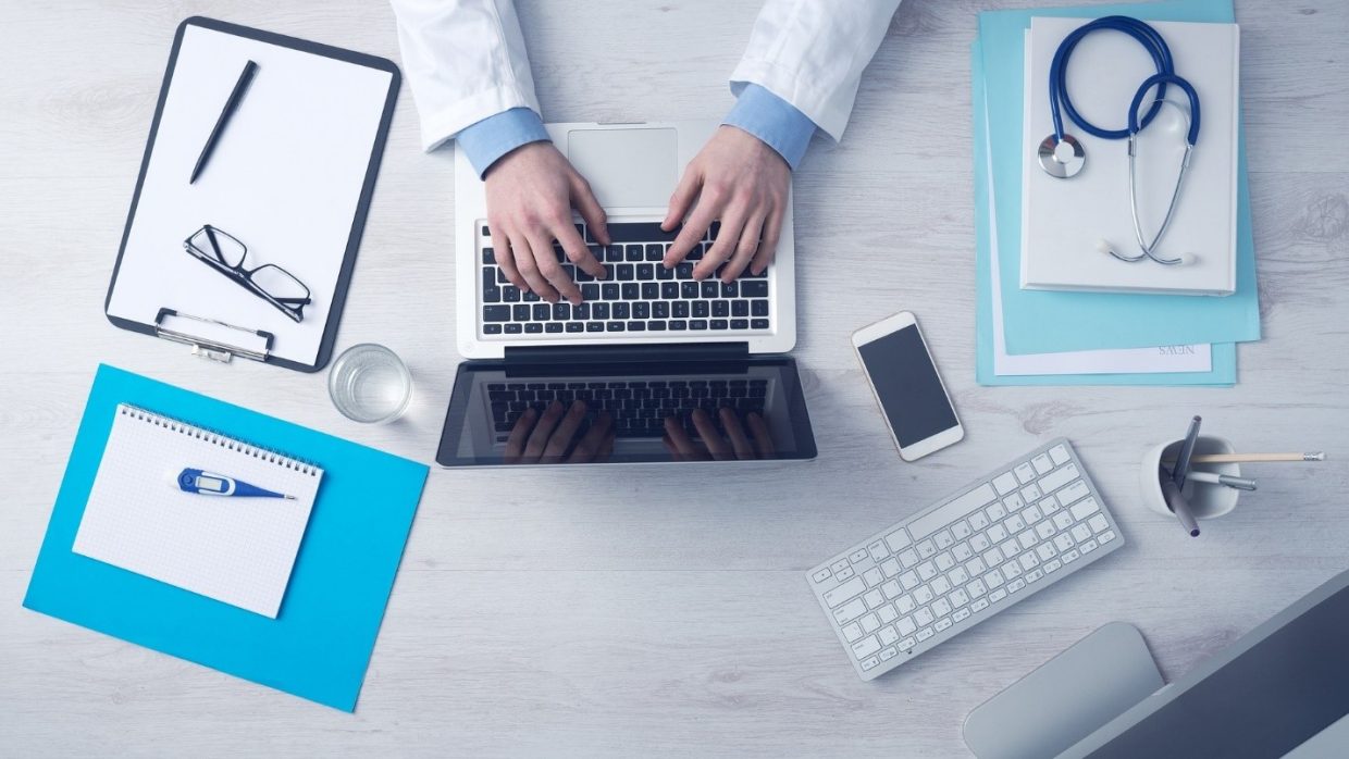 A clipboard with a pen and glasses on it, a notebook with a thermometer, a glass of water, a phone, a spare keyboard, a few files with a stethoscope on top of them, a pen holder and a medical professional's hands shown working on a laptop