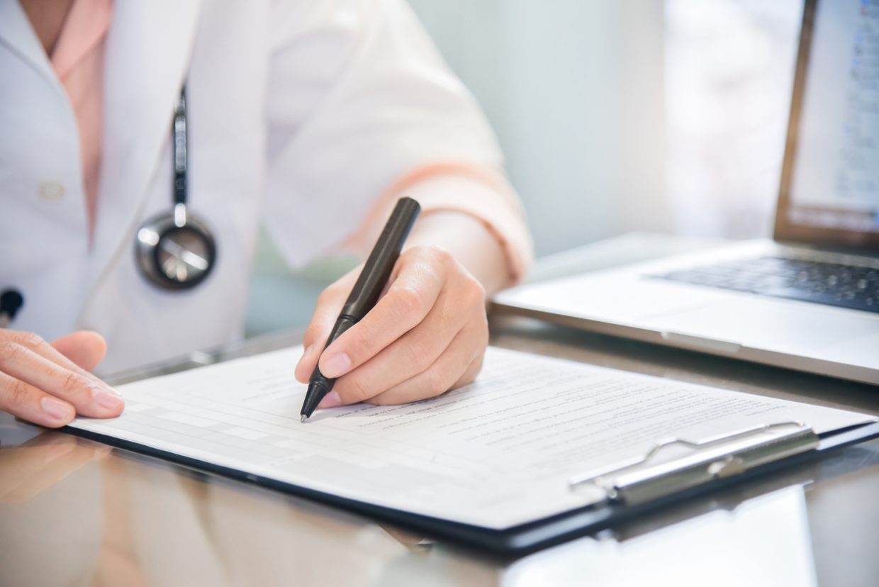 Person in scrub coat with stethoscope around neck is writing something on a clipboard. There is a laptop open beside the clipboard.