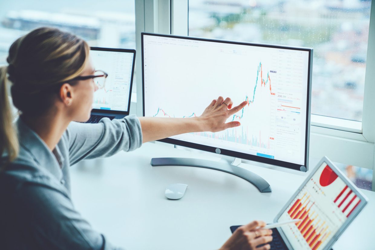 A woman studying a graph on a monitor, with a laptop and an iPad nearby.