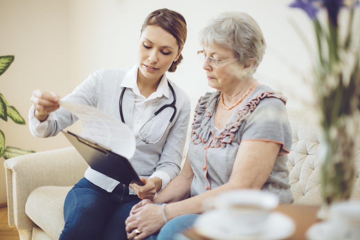 Medical professional with a stethoscope around her neck is consulting with a senior lady on a sofa. As she speaks, she is showing and sharing the examination results to the lady.