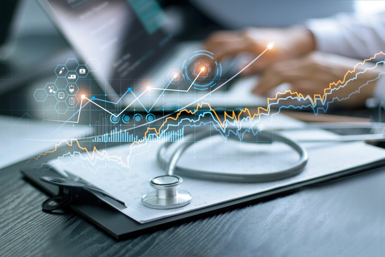 A graph representing growth is overlaid on top of a clipboard with a stethoscope. In the background, a person is working on a laptop.