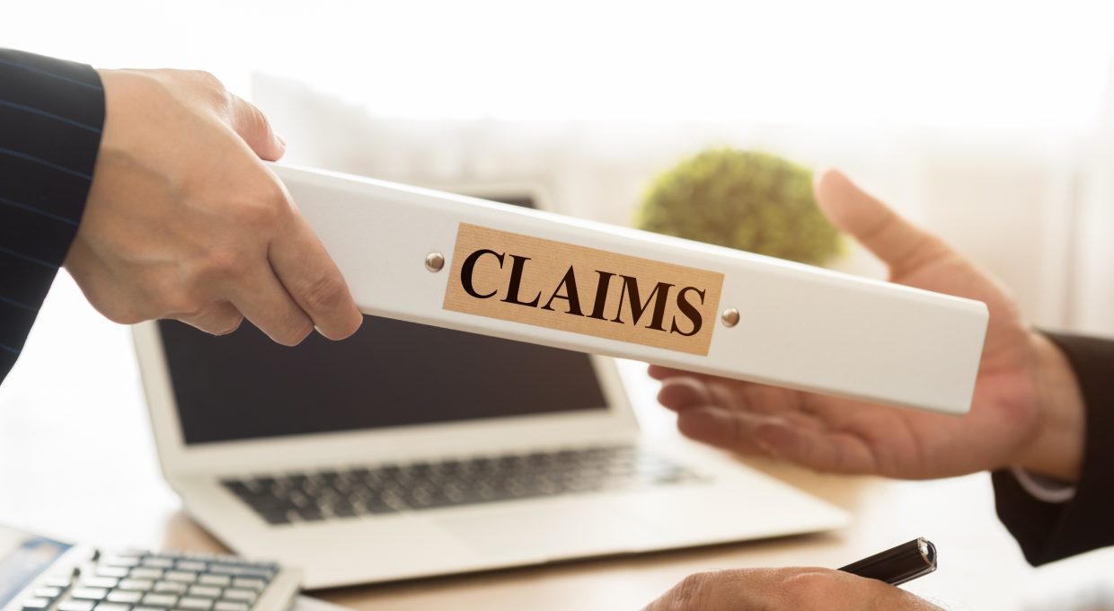 A person handing over a file labeled claims to another person. A laptop is visible in the background.
