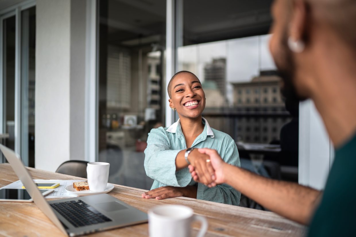 A woman sitting on a table with a laptop in front of her, along with 2 phones, a few papers, and a mug of coffee with cake. She is shaking hands with a man who also has a mug of coffee in front of him.