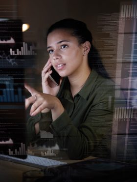 A woman on a phone call analyzing data charts and code overlays on a computer screen at night.